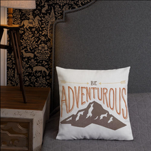 Load image into Gallery viewer, A pillow leaning on a grey bed headboard. The white pillow features the phrase “Be adventurous” with arrows pointing to the word “be” and a brown mountain illustration underneath the word “adventure.”
