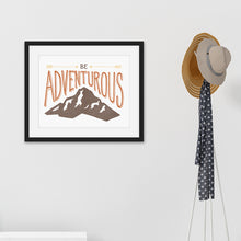 Load image into Gallery viewer, Black framed artwork hanging on a wall next to a coat rack. The lettering and illustration says “Be adventurous” with arrows pointing to the word “be” and a brown mountain illustration underneath the word “adventure.”