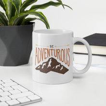 Load image into Gallery viewer, A mug featured on a desk with a plant and a keyboard. The white mug features the phrase “Be adventurous” with arrows pointing to the word “be” and a brown mountain illustration underneath the word “adventure.”