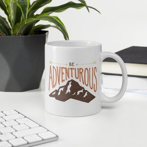 A mug featured on a desk with a plant and a keyboard. The white mug features the phrase “Be adventurous” with arrows pointing to the word “be” and a brown mountain illustration underneath the word “adventure.”
