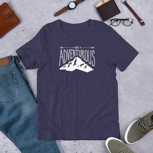 A short sleeved T-shirt laying flat with objects around it. The tee is in the color heather midnight and features the lettering and illustration in white. The phrase “Be adventurous” with arrows pointing to the word “be” and a mountain illustration underneath the word “adventure.”