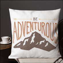Load image into Gallery viewer, A pillow on a chair with a coffee mug on a table next to it. The white pillow features the phrase “Be adventurous” with arrows pointing to the word “be” and a brown mountain illustration underneath the word “adventure.”