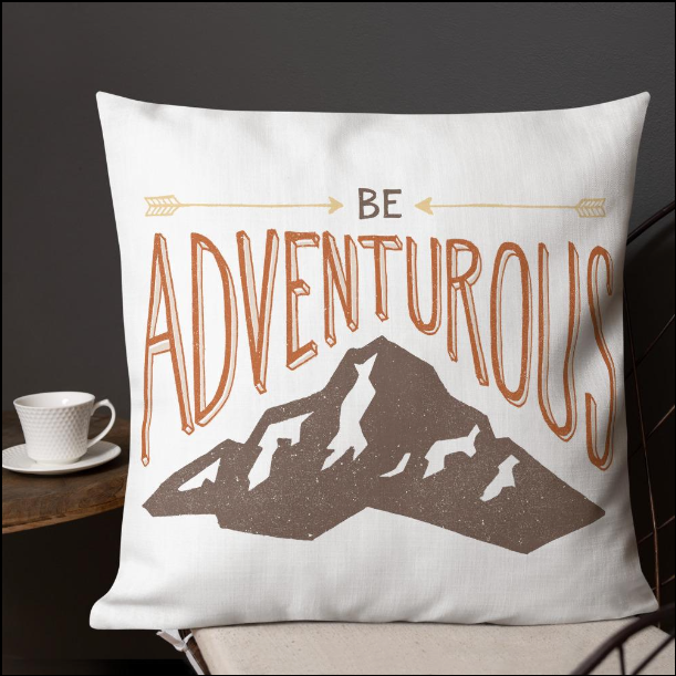 A pillow on a chair with a coffee mug on a table next to it. The white pillow features the phrase “Be adventurous” with arrows pointing to the word “be” and a brown mountain illustration underneath the word “adventure.”