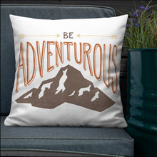 Load image into Gallery viewer, A pillow leaning on a grey sofa with a plant in the background. The white pillow features the phrase “Be adventurous” with arrows pointing to the word “be” and a brown mountain illustration underneath the word “adventure.”