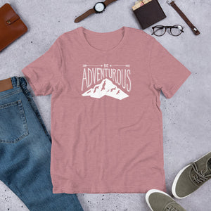 An orchid pink T-shirt laying flat with objects around it. The tee features the lettering and illustration in white. The phrase “Be adventurous” with arrows pointing to the word “be” and a mountain illustration underneath the word “adventure.”