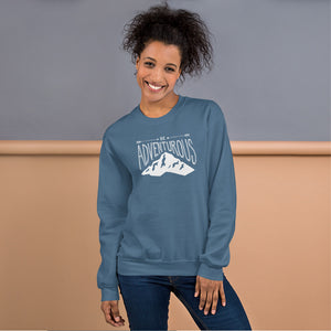 A woman wearing an indigo blue hoodie with lettering and illustration in white with the phrase “Be Adventurous” with arrows pointing to the word “be” and a mountain illustration underneath the word “adventure.”
