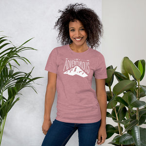 A woman wearing an orchid pink short sleeved t-shirt. The tee features the lettering and illustration in white. The phrase “Be adventurous” with arrows pointing to the word “be” and a mountain illustration underneath the word “adventure.”