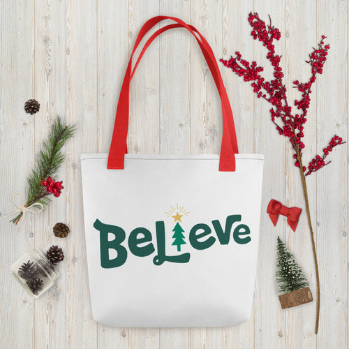 A white tote bag with red handles laying on a table with Christmas items around it. The tote bag features the word Believe in green with an illustrated Christmas tree as the 