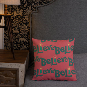 A pillow leaning on a grey headboard with a table and lamp off to the side. The white pillow is red with the word Believe in green and the "I" of the word as an illustrated Christmas tree. The pattern repeats on the pilow. 