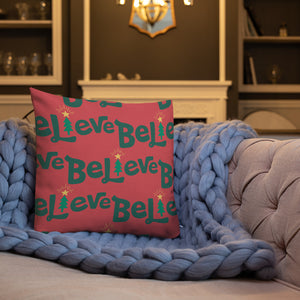 A white pillow on a sofa with a blue knitted blanket. The red pillow features the word Believe with the "I" as an illustrated Christmas tree. The words are in green on the red pillow. The pattern repeats over the pillow. 