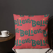 Load image into Gallery viewer, A pillow on a chair with a coffee mug on a table next to it. The red pillow features the word Believe with the &quot;I&quot; as an illustrated Christmas tree. The pattern repeats over the pillow. 