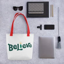 Load image into Gallery viewer, A tote bag lying on a surface with a laptop and office items next to it. The tote bag has the word Believe in the center. The &quot;I&quot; of the word Believe is an illustrated Christmas tree. The handles are red. 