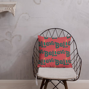 The pillow is leaning on a metal chair with a cushion. The red pillow features the word Believe in green with the "I" of the word as an illustrated Christmas tree. 
