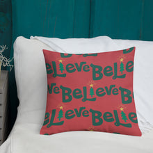 Load image into Gallery viewer, A white pillow with illustrations leading on white bedding with a side table off to the side. The red pillow features the word Believe in a repeat pattern with the word in green. The &quot;I&quot; of the word Believe is featured as an illustrated Christmas tree. 