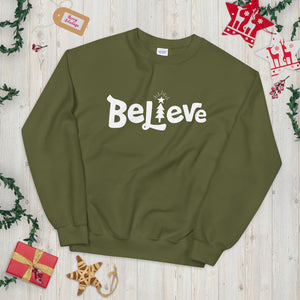 A hunter green sweatshirt laying on a table with Christmas objects around it. The sweatshirt features the word Believe in white with an illustrated Christmas tree as the "I."