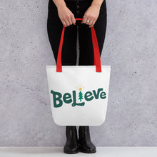 Load image into Gallery viewer, Someone holding a tote bag with red handles and a white fabric bag. The artwork is the word Believe with an illustrated Christmas tree as the &quot;I.&quot; The word is in green in the center of the bag. 