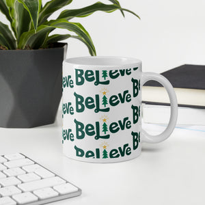 A mug featured on a desk with a plant and a keyboard. The white mug has the word "Believe" in green. The "I" of Believe is an illustrated Christmas tree. The word is in a repeat pattern on the mug. 