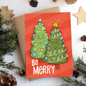 A photo of a Christmas card on top of a brown paper wrapped gift with Christmas decor around it. The Christmas card has a red background with two illustrated cute Christmas trees and the words "Be Merry" in white. 