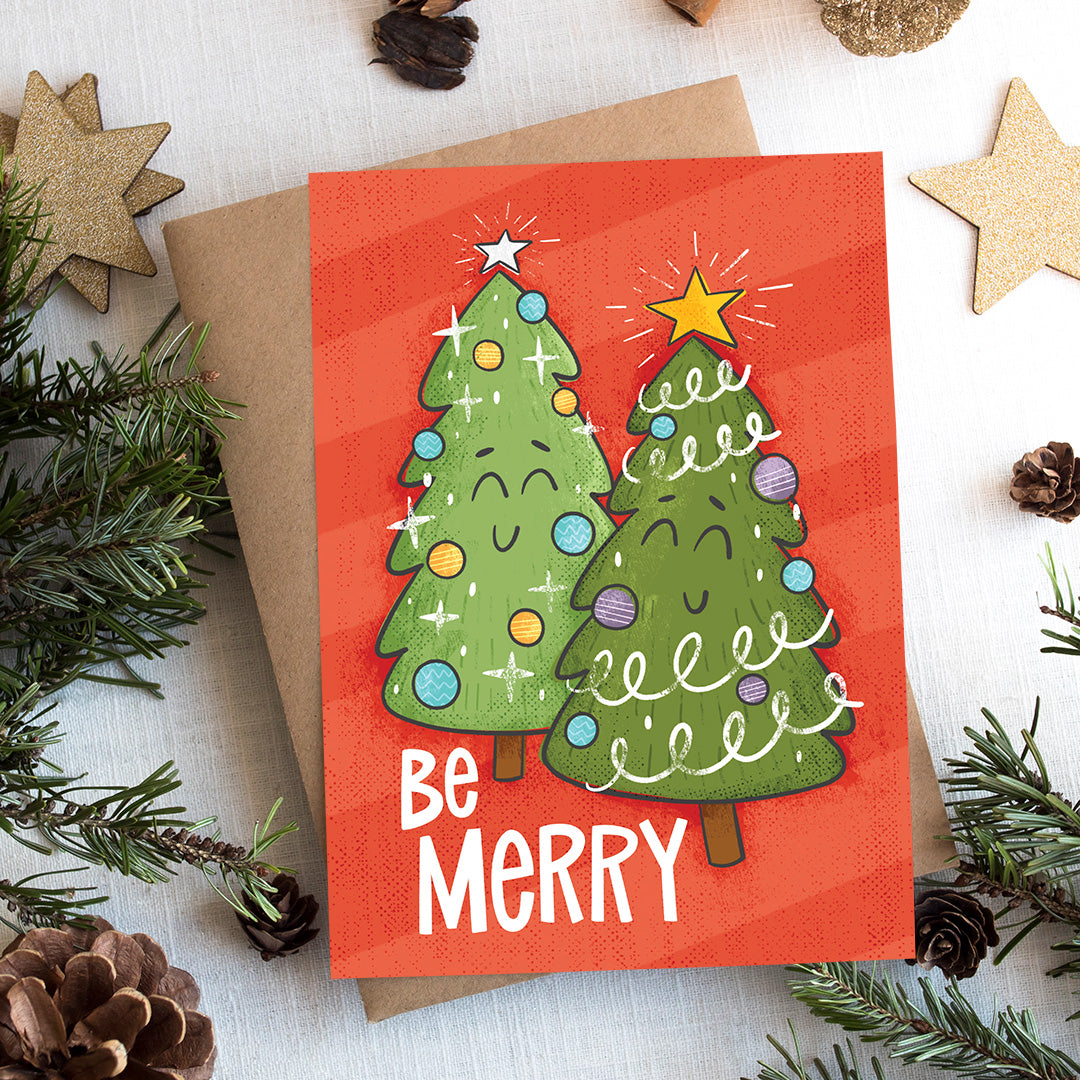 A photo of a Christmas card on top of a brown paper wrapped gift with Christmas decor around it. The Christmas card has a red background with two illustrated cute Christmas trees and the words 