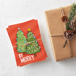 A stack of Christmas cards with brown string wrapped around them. A brown craft paper gift is off to the side. The Christmas card has a red background with two illustrated cute Christmas trees and the words "Be Merry" in white.