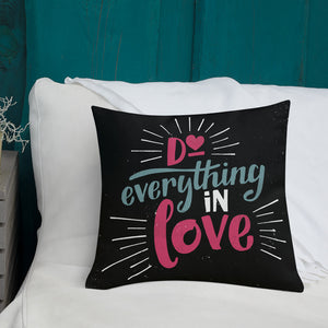 A square black pillow sits on a white sofa. The pillow reads "Do everything in love" in bright pink and blue hand-lettering style, with white dashes around the words.