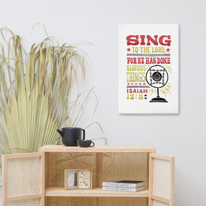 A canvas shown on a wall above a shelf. The canvas has a white background with the words "Sing to the Lord for he has done glorious things. Isaiah 12:5." The words are in red, yellow and black. 