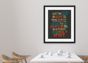 Artwork in a black frame with the with a white matte hanging on a wall in a dining room. The artwork is on a black background with colorful letters reading May the God of hope fill you with all joy and peace as you trust him, so that you may overflow with hope by the power of the holy spirit." 