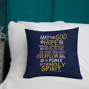 A pillow on a bed with the Bible verse "May the God of hope fill you with all joy and peace as you trust him, so that you may overflow with hope by the power of the holy spirit." The pillow is purple with lettering in white, pink and yellow. 