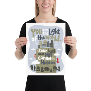 A woman holding a white piece of paper with artwork on it. The artwork is on a white background with lettering reading "You are the light of the world, a town built on a hill cannot be hidden." The words are a light gray background with an illustrated city.