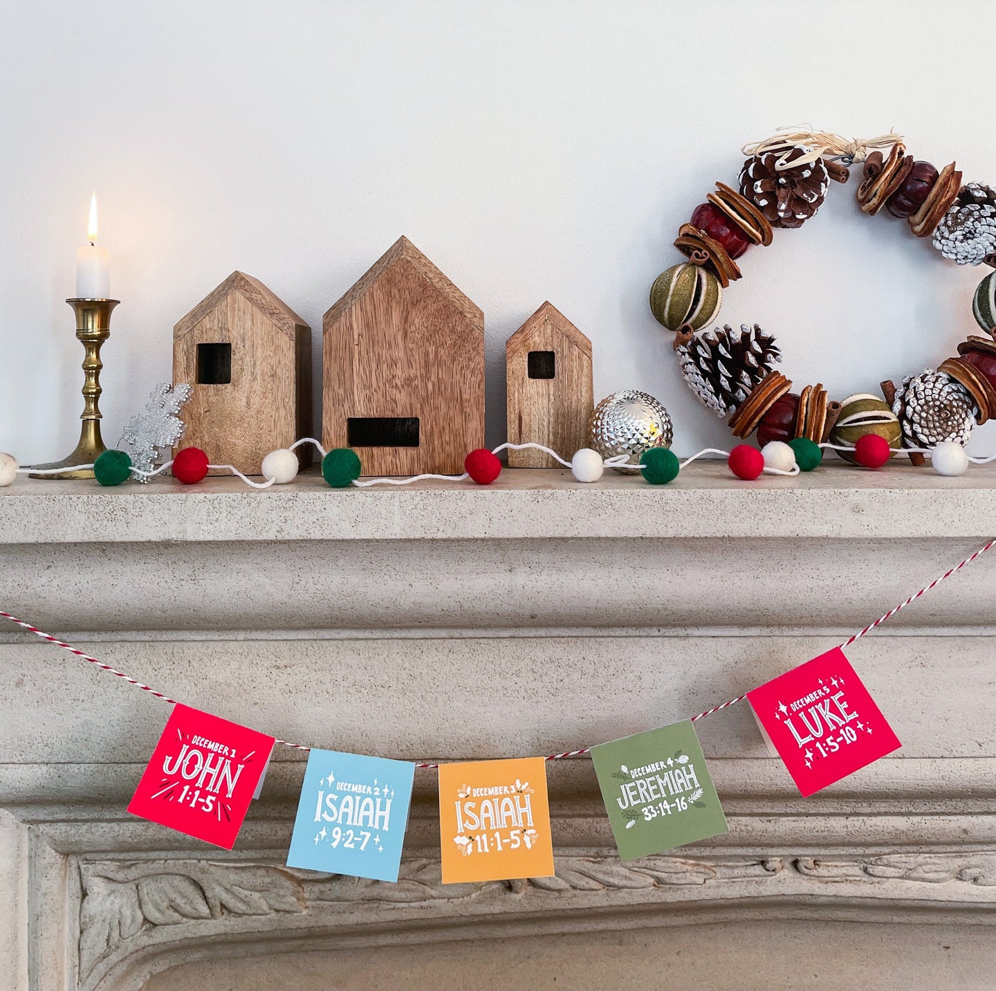 A picture of a fireplace mantle with a candle lit, small houses and a Christmas wreath. Hanging on the mantle on a string are the advent calendar cards showing the Bible verses. 