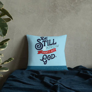 A bright blue cushion sits on a dark blue bed against a grey concrete wall. The cushion features the verse 'Be Still and Know that I am God' illustrated in a bold typographic style.