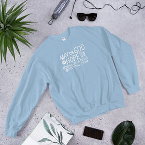 A light blue sweatshirt laying on the ground with objects around it. The sweatshirt features hand drawn lettering in white with the words "May the God of hope fill you with all joy and peace as you trust him."