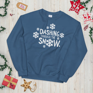 A indigo blue sweatshirt laying on a table with Christmas objects around it. The sweatshirt has the words "Dashing through the snow" in white. There are snowflakes around the letters. 