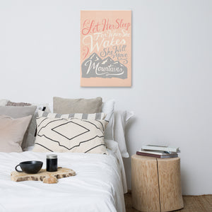 An illustrated pink canvas hangs on a white wall, over a white bed with lots of cushions and pillows. The canvas reads 'Let her sleep for when she wakes she will move mountains' in a pink, white, and light grey lettering design, with a grey mountain illustration at the bottom.
