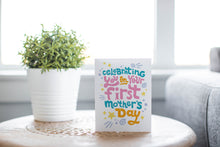Load image into Gallery viewer, A photo of a card featured on a tabletop next to a white planter filled with a green plant. ​​The card features the words “Celebrating you on your first Mother’s Day.”