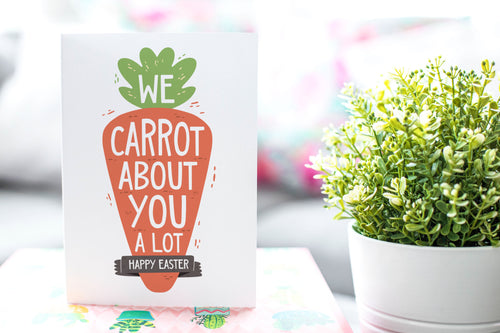 A photo of a card featured on a tabletop next to a white planter filled with a green plant. ​​The card features the words “We carrot about you a lot, Happy Easter,” all featured in an illustrated carrot.