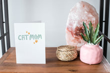 Load image into Gallery viewer, A card on a wood tabletop and on the right side of the card is a woven basket, a pink plant pot with a cactus in it and a pink crystal rock. The card features the words “Cat Mom.”