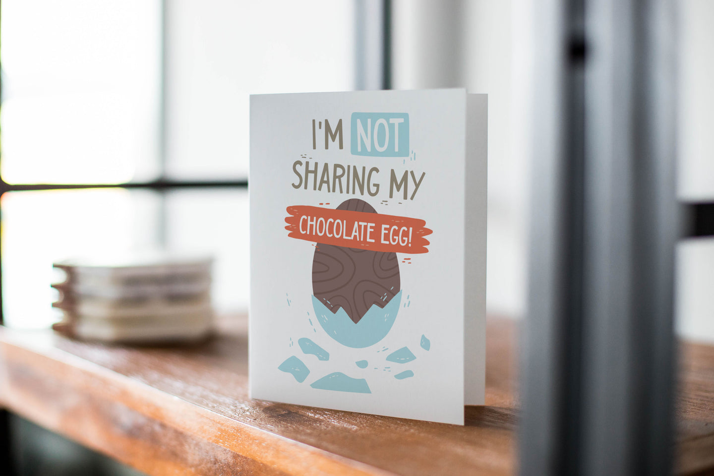A card on a wood tabletop with an object in the background that is out of focus. The card features an illustrated chocolate Easter egg with the words “I’m not sharing my chocolate egg!”
