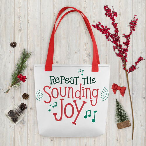 A white tote bag with red handles laying on a table with Christmas items around it. The tote bag features the words 
