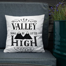 Load image into Gallery viewer, A monochrome square pillow sits on a grey outdoor sofa against a dark wooden fence. The pillow design is black on a white background, and reads &#39;Every valley shall be lifted high&#39; in a variety of typographic lettering, with flourishes and an illustration of two mountain peaks. Next to the soda is a metal container filled with purple flowers. 