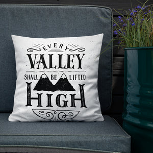 A monochrome square pillow sits on a grey outdoor sofa against a dark wooden fence. The pillow design is black on a white background, and reads 'Every valley shall be lifted high' in a variety of typographic lettering, with flourishes and an illustration of two mountain peaks. Next to the soda is a metal container filled with purple flowers. 