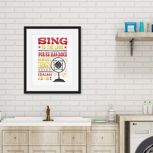 Artwork featured on a kitchen wall with a black frame. The artwork is on a white background with lettering reading "Sing to the Lord for he has done glorious things. Isaiah 12:5." The words are in red, yellow and black. 