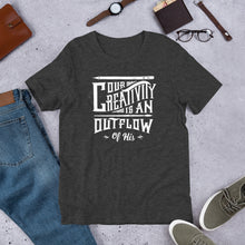 Load image into Gallery viewer, A short sleeved T-shirt laying flat with objects around it. The tee is a dark grey heather color and features hand drawn lettering in white with the words &quot;Our creativity is an outflow of His.&quot;