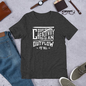 A short sleeved T-shirt laying flat with objects around it. The tee is a dark grey heather color and features hand drawn lettering in white with the words "Our creativity is an outflow of His."