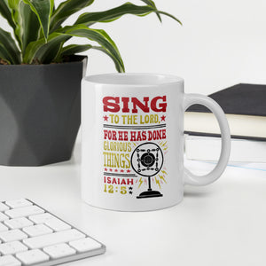 A mug featured on a desk with a plant and a keyboard. The white mug features hand drawn lettering with the words "Sing to the Lord for he has done glorious things. Isaiah 12:5." The words are in red, yellow and black.