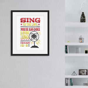 Artwork featured on a wall in a black frame by a shelving unit. The artwork is on white paper and features hand drawn lettering with the words "Sing to the Lord for he has done glorious things. Isaiah 12:5." The words are in red, yellow and black. 