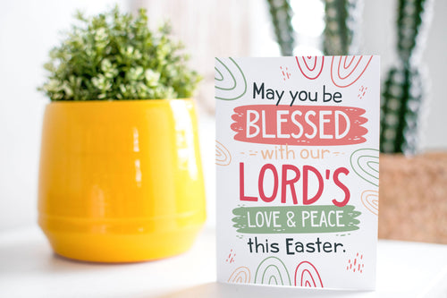 A greeting card is on a table top with a yellow plant pot and a green plant inside. The card features the words “May You be Blessed with our Lord's Love & Peace this Easter.”