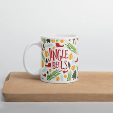 Load image into Gallery viewer, A white mug sitting on a light wood cutting board. The white mug shows the red words &quot;Jingle Bells&quot; in the middle of Christmas themed illustrations. The illustrations are sleighs, pine trees, leaves, and ornaments. The illustrations are in the colors light and dark green, light blue and yellow. 
