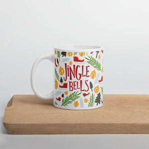 A white mug sitting on a light wood cutting board. The white mug shows the red words "Jingle Bells" in the middle of Christmas themed illustrations. The illustrations are sleighs, pine trees, leaves, and ornaments. The illustrations are in the colors light and dark green, light blue and yellow. 