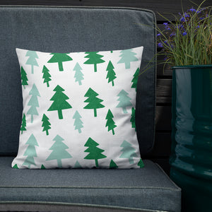 The white pillow is leaning on a sofa with a plant off to the side. The pillow is white with illustrations in the colors dark and light green. The illustrations are of pine trees. 